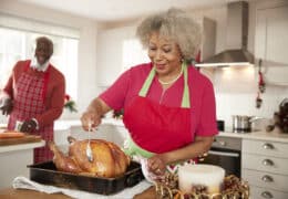 Senior black woman basting a roast turkey in preparation for Christmas dinner, her husband chopping vegetables in the background, front view, close up